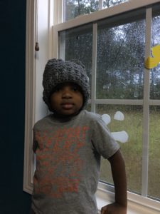 A young male boy standing in front of a window on a rainy day. He is wearing a gray, slouchy crochet hat.