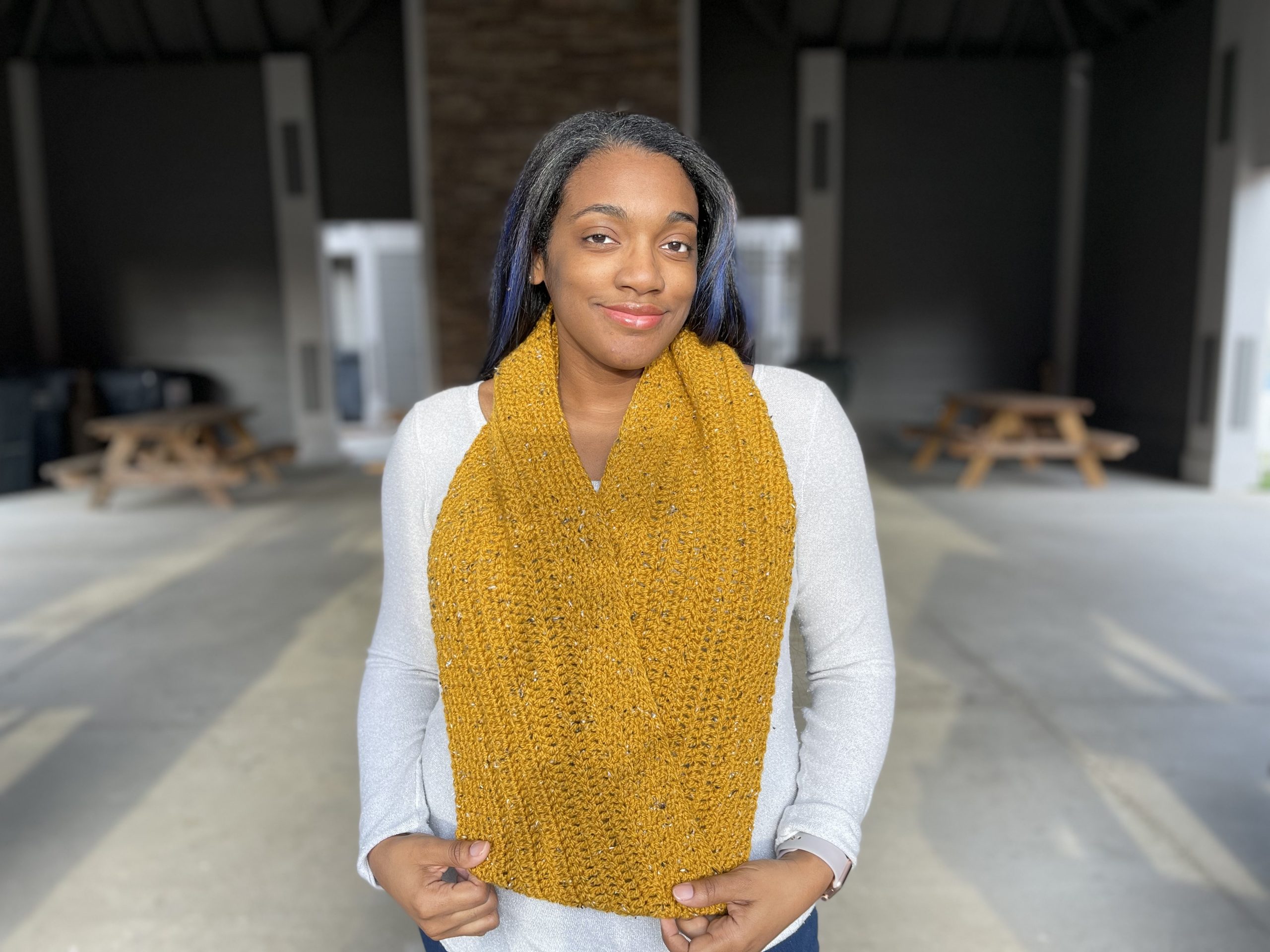 An image of an infinity crochet scarf made with a subtle texture in a mustard tweed color yarn. The woman is smiling while wearing this crochet scarf.