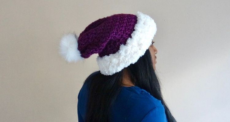 A picture of a royal purple crochet hat with a white faux fur pom and white faux fur edging on a woman with long, black hair.