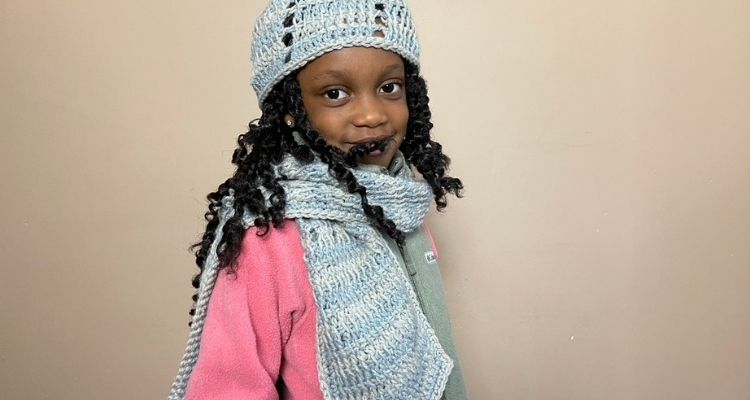 An image of a female child wearing a striped tunisian crochet scarf and tunisian crochet hat. The yarn for the crochet scarf and hat is a dusty blue and light gray color.