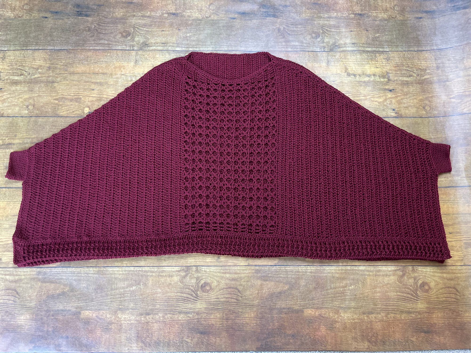 Image shows a burgundy trapezoid shaped crochet garment with small wrist cuffs on each side. The center is created with an open stitch, and it has a ribbed hem.