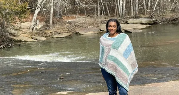 An image of a Black woman wearing a white, muted green, and two shades of soft brown striped tunisian crochet poncho. There is a stream flowing behind her on a sunny day.