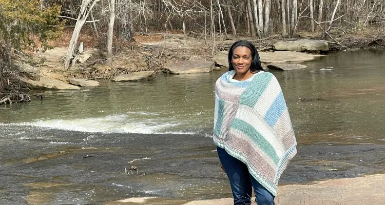 An image of a Black woman wearing a white, muted green, and two shades of soft brown striped tunisian crochet poncho. There is a stream flowing behind her on a sunny day.