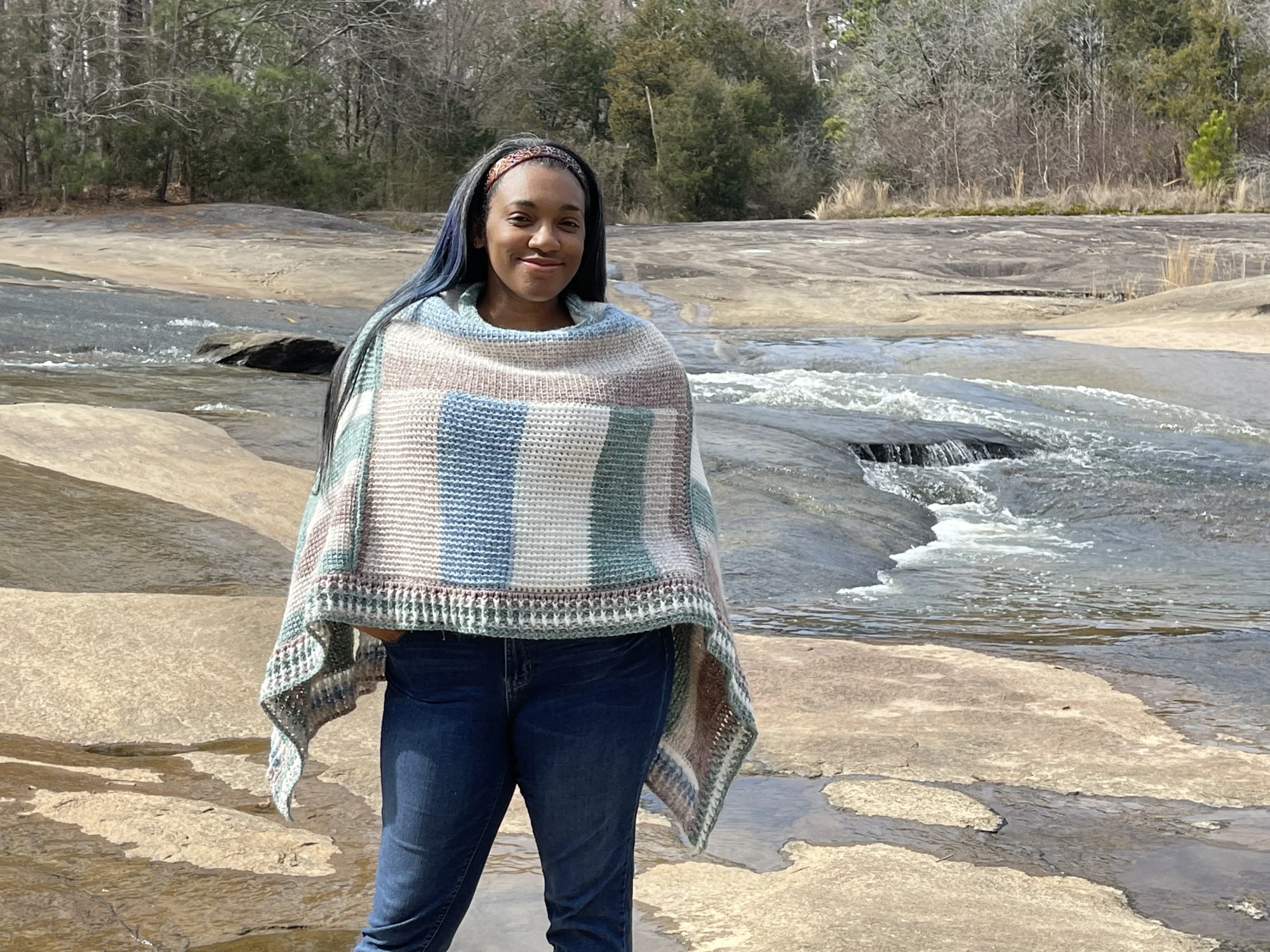 A black woman is wearing a cream, green, blue, tan, and brown stripe tunisian crochet poncho. She is standing  with streams of water flowing over smooth rock in the background. 