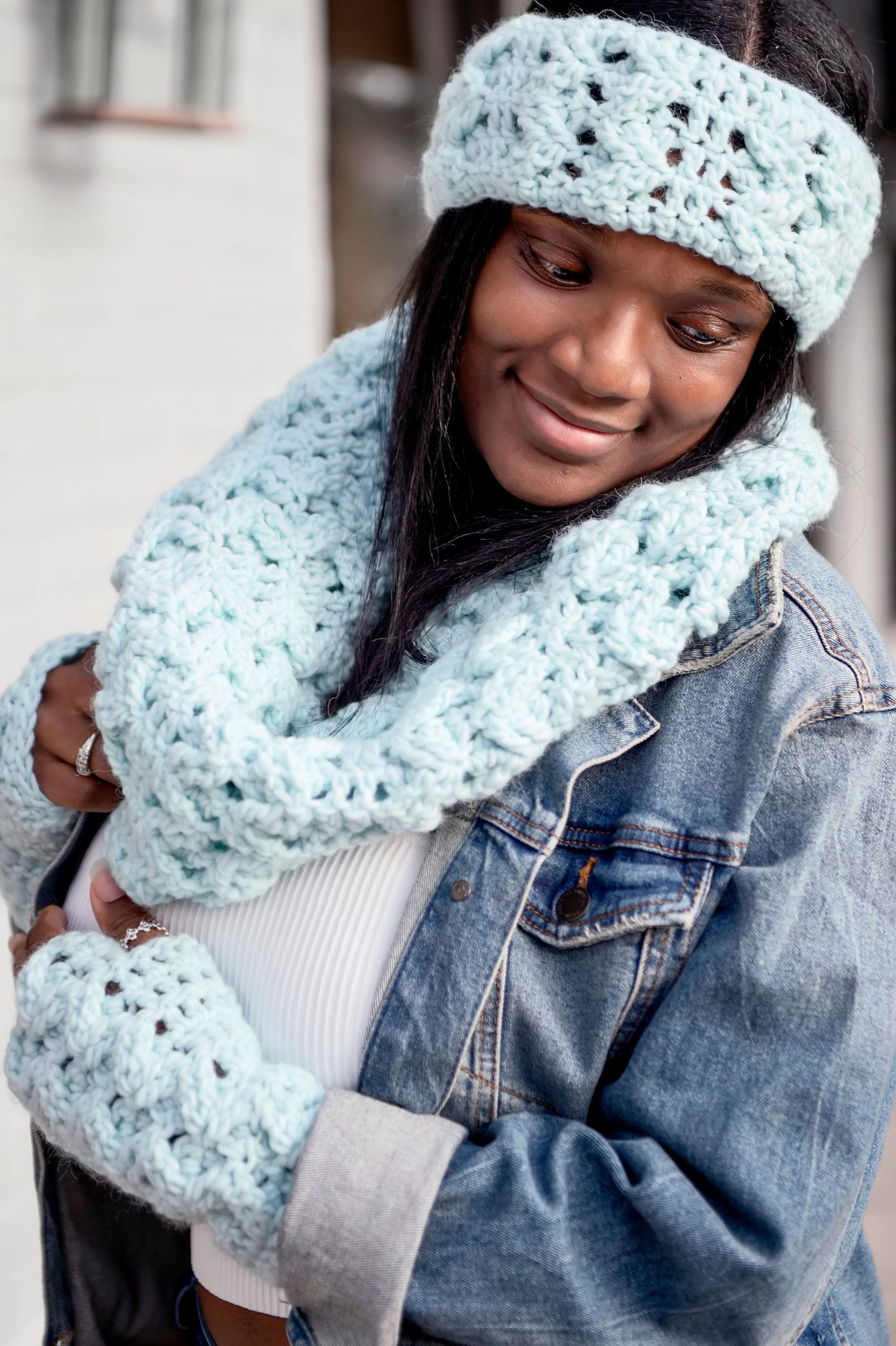 A woman looking downward and smiling while wearing a seafoam colored and textured crochet ear warmer, crochet cowl and crochet fingerless mitts