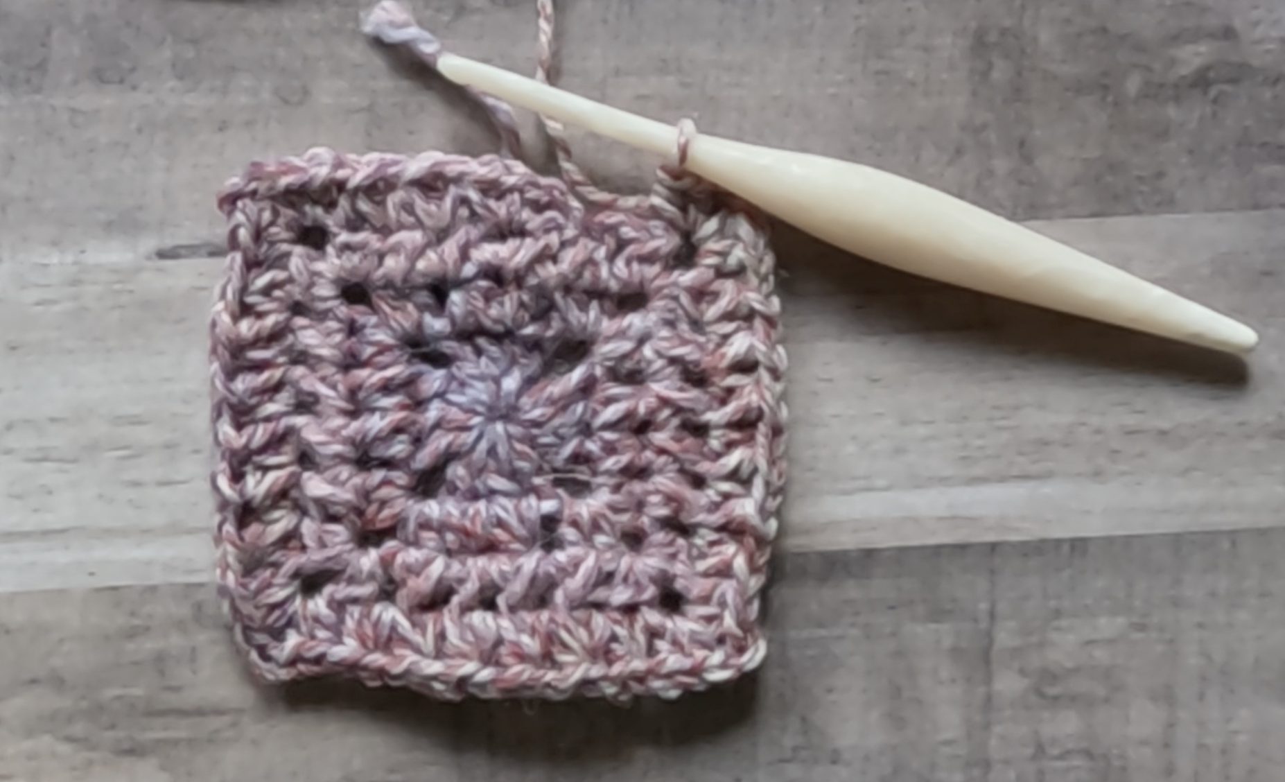 An image of a crochet granny square being made with a purple color yarn and cream colored crochet hook. The crochet square shows 3 completed rounds and the 4th round is almost complete.