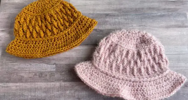 Two crochet hats featuring a textured stitches. One is a soft pink, and the other is a golden yellow, and appears smaller in size.