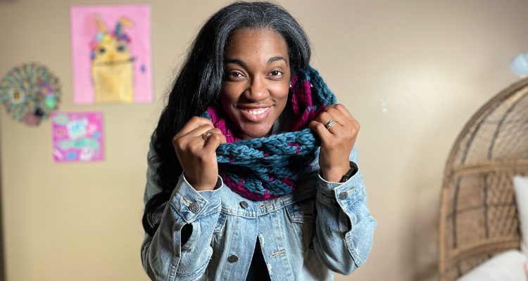An image of a woman smiling and holding onto a chunky crochet cowl that is a dark teal on the outside and peaks of magenta on the inside.