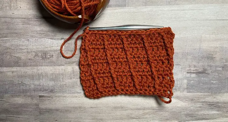 Raised Diagonal Crochet Tutorial image of burnt orange yarn in a rectangle swatch of double crochet stitches and diagonal front post stitches, a yarn bowl and silver metal crochet hook.