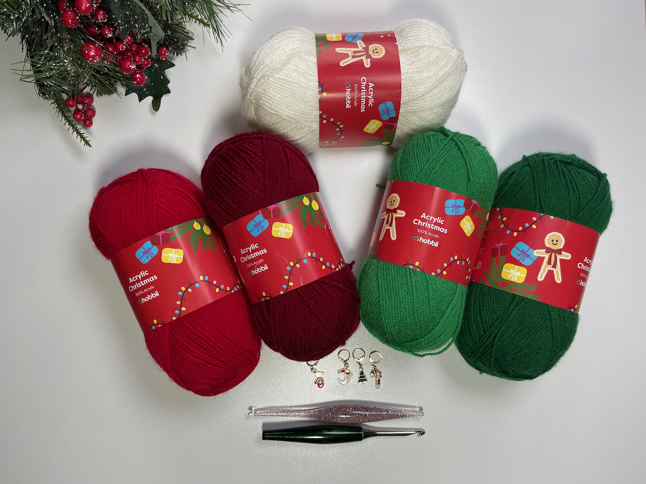 Hobbii Christmas Acrylic Color Pack yarn in two shades of green and two shades of red and cream with a glitter crochet hook, an emerald crochet hook, and four Christmas stitch markers that will be used to make the Christmas Crochet capelet.