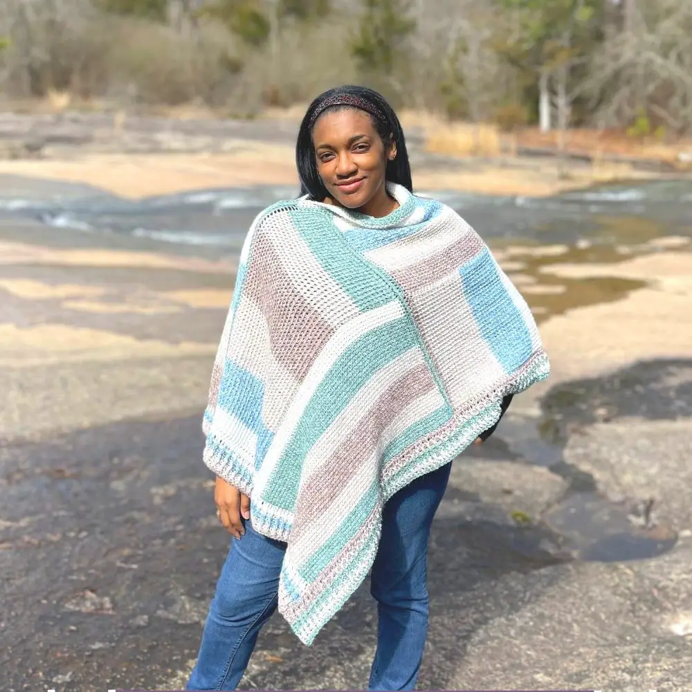 Image of a African American woman wearing a striped poncho. The stripes are green, beige, brown and green in color, and go in vertical and horizontal directions.