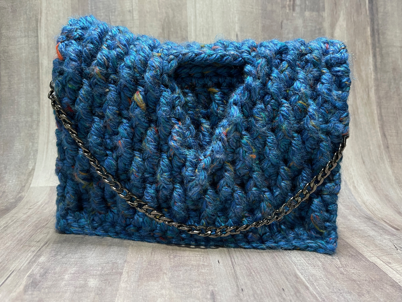 Image of a a crochet bag in chunky blue yarn. The bag is made with textured post stitches and a triangle opening. This crochet pattern is only available in the Crochet for Me bundle.