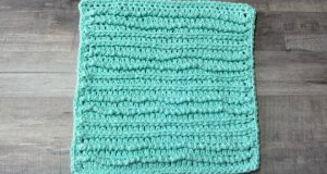 An image of a textured crochet dishcloth in a turquoise cotton yarn. There are rows of stitches on the surface of the cloth made from reverse single crochet and yarn over slip stitches.