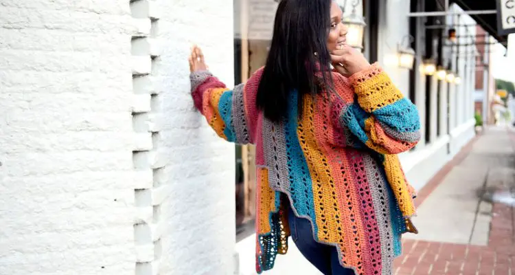 The back of the Xcapade Cardigan is shown demonstrating the striping effect and stair step hemline. The model has her left hadn't against a white brick wall with her head turned towards her right shoulder and her right hand on her cheek.