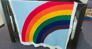 An image of a tunisian crochet blanket with a curving rainbow rising from the lower left side to the right side. The background is a sky blue and the edging is white with tunisian bobble stitches to represent clouds. The tunisian crochet blanket is draped on the side of a crib.