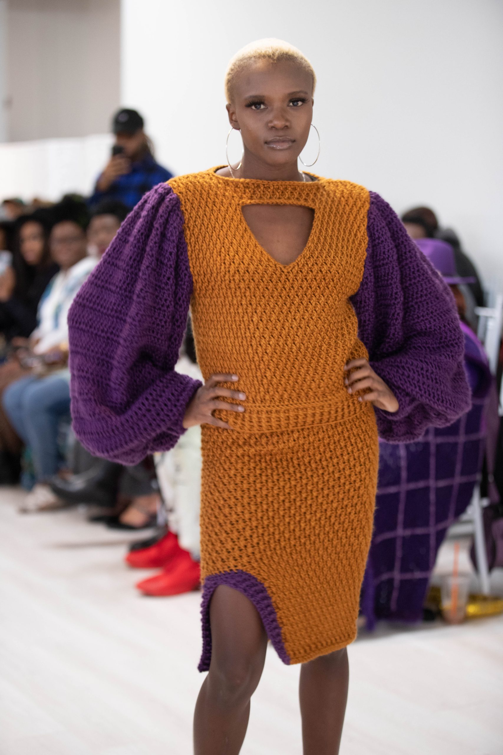 An image of an African American woman with short blonde hair wearing a crochet sweater and skirt. The crochet sweater has a triangle opening above the breasts, purple balloon sleeves and the body is a golden caramel color. The Skirt matches in color, but has a split on the right leg that is lined in purple ribbing.