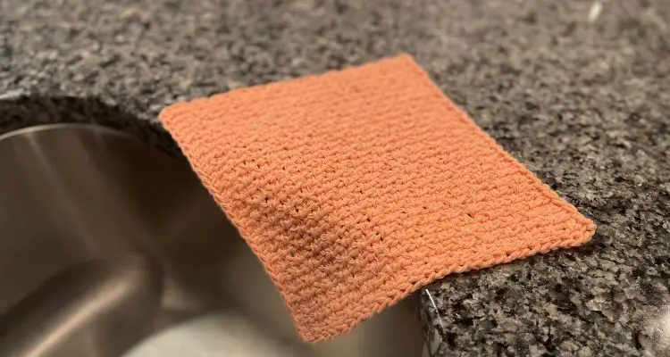 Wide image for a blog post of a peach colored tunisian crochet dishcloth laying on the edge of a black and gray granite countertop and hanging down into a stainless steel sink.