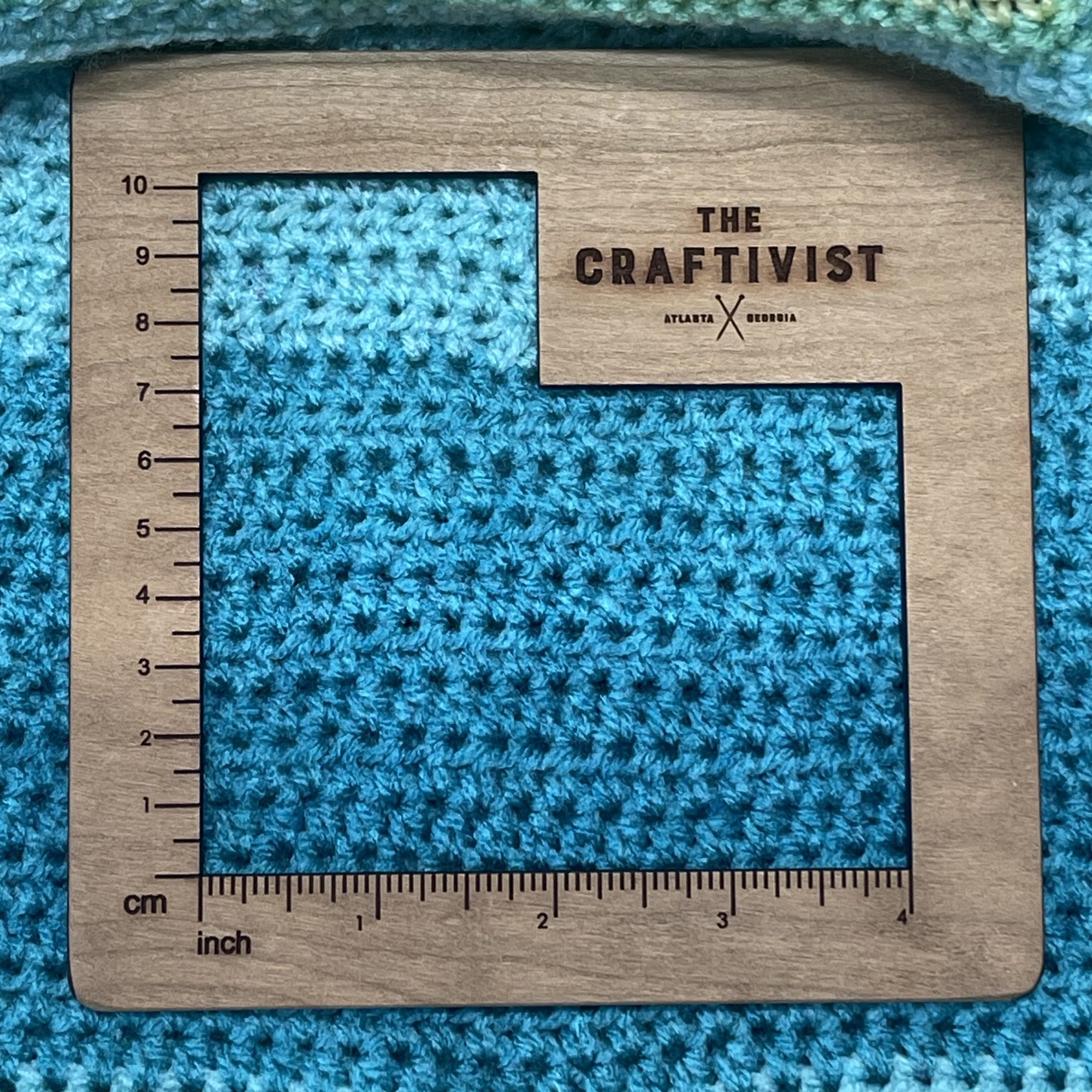 A wood gauge square with inches indicated on the lower side and centimeters listed on the left side. It is laying on top of a crochet fabric made with turquoise yarn to show crochet gauge.