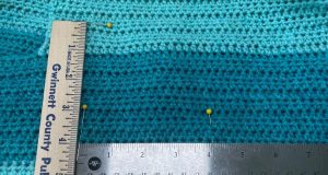 An image of a stainless steel ruler laying horizontally and a wood ruler laying vertically with there yellow straight pins in a turquoise crochet fabric showing crochet gauge being measured.