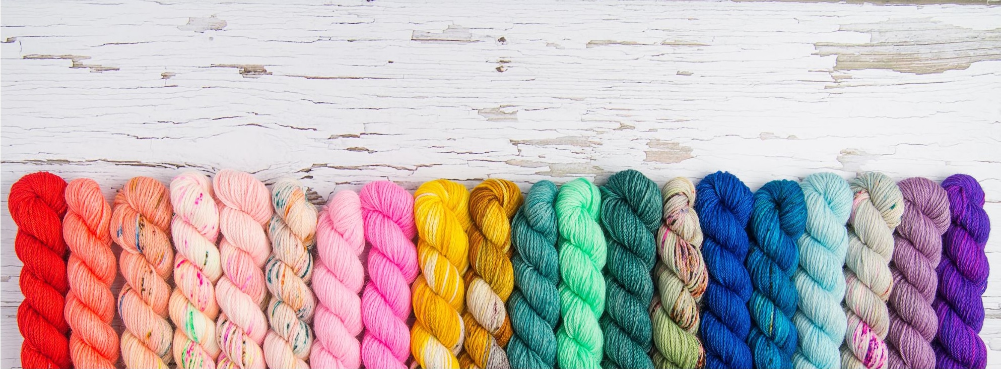 An image of 20 hanks of yarn on a piece of whitewashed wood. From left to right, the yarn colors range from red to coral to shades of pink with speckles of other color, two shades of yellow, shade of teal to a mint green to a darker heather green to a variegated with green and brown to blues and ends with a tonal purple. This is in reference to choosing yarn for crochet bottoms.
