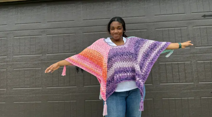 A Black woman is wearing a colorful crochet poncho showing two colors in the granny stitch. You can see 4 of the 6 points and tassels of this hexagon crochet poncho.