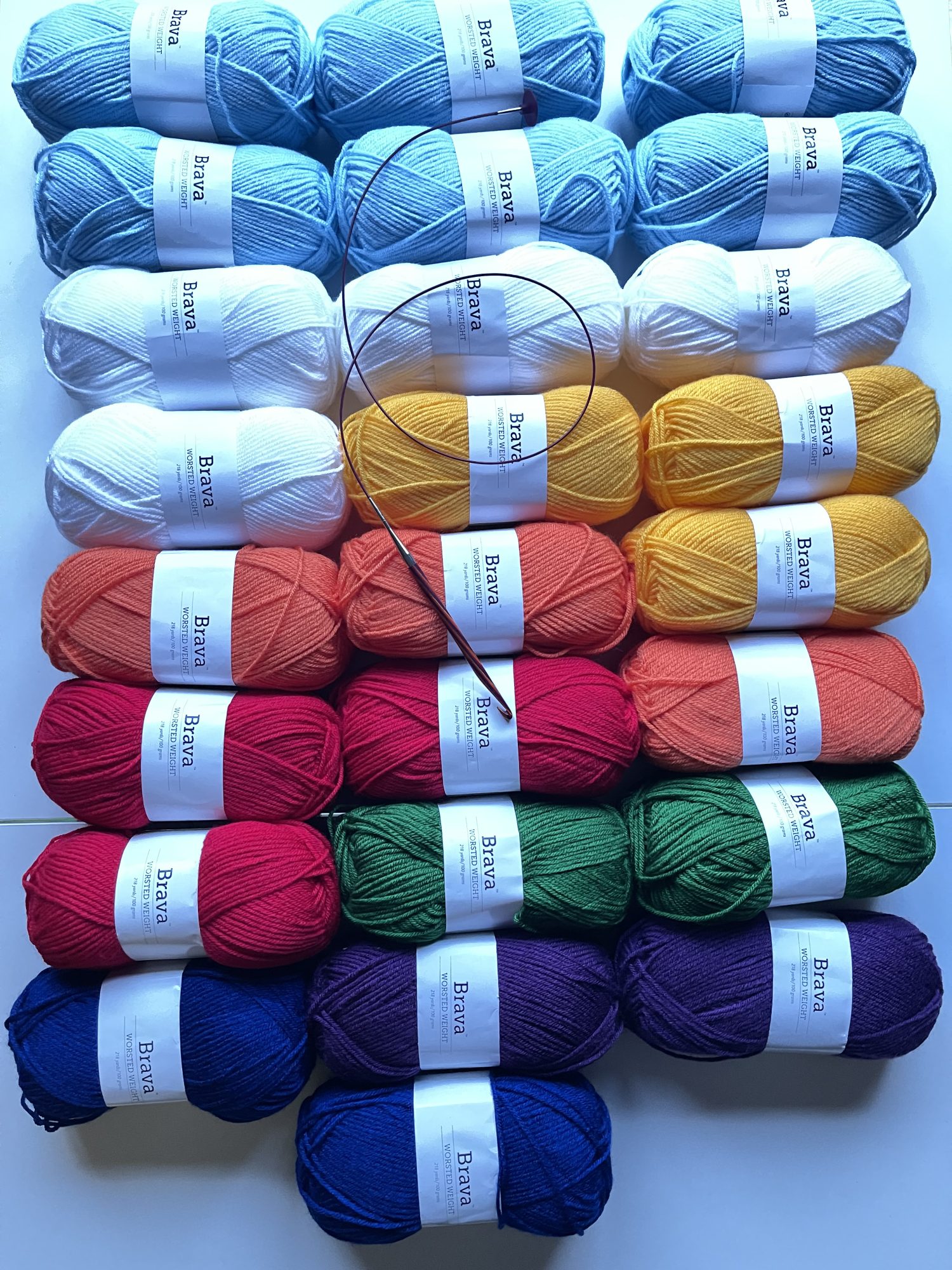 An image of yarn that is grouped by color for a tunisian blanket. Starting at the top, there are 6 skeins of sky blue yarn, 4 skeins of white yarn, 3 skeins each of yellow, orange and red, and 2 skeins each of green, purple and blue. There is a multicolored tunisian crochet hook with an attached cable laying on top of the yarn for the tunisian blanket.