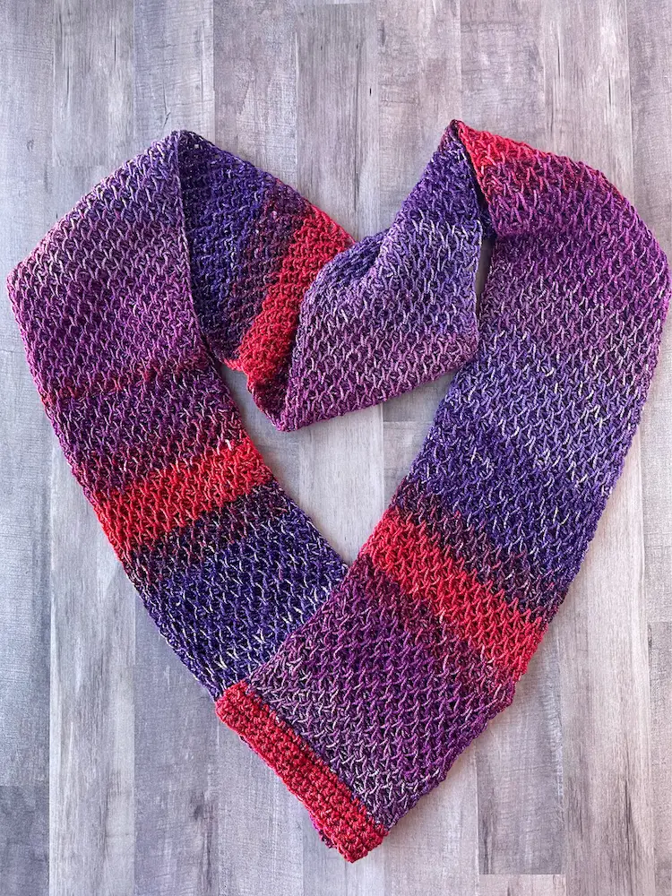 An image of a tunisian crochet scarf made in the tunisian smock stitch. The tunisian crochet scarf is made with  a yarn that changes from red to purples to burgundy.