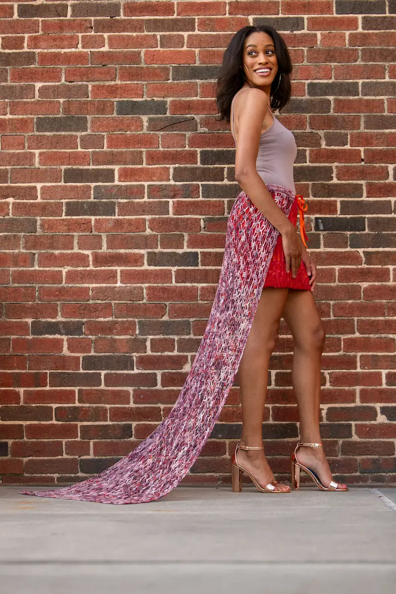 An African American woman smiling while wearing a red-orange crochet mini skirt with a variegated lace train and standing in front of a red brick wall. 