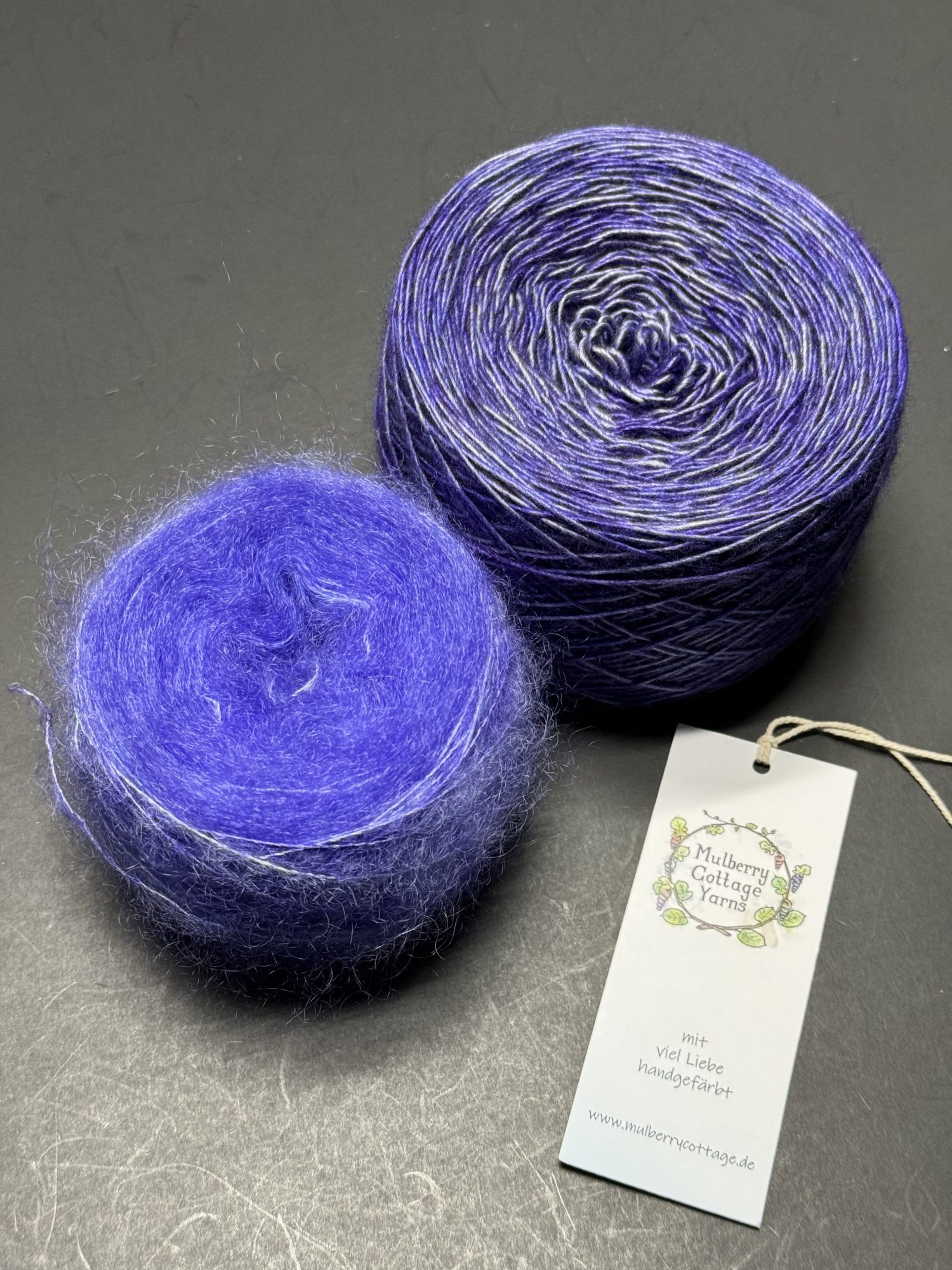 An image of two cakes of yarn. The yarn cake on the lower left appears fuzzy and very thin in a purplish blue color. The yarn cake in the upper right is the same color but slightly thicker and a single strand.