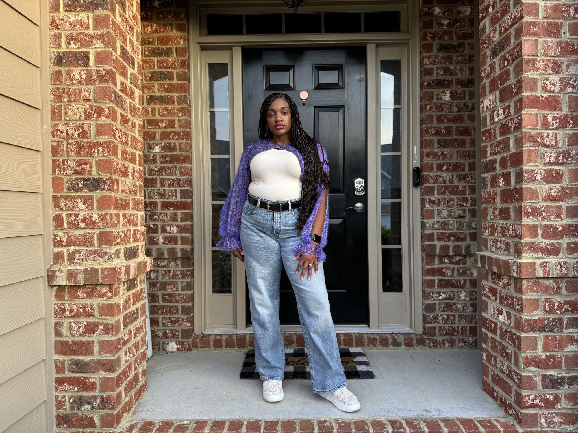 An image of an African American woman standing in front of a black door with side windows wearing a crochet bolero. The crochet shrug is a purple color, cropped length, closes in the middle at the neckline, and becomes narrow towards the hem. The sleeves are made with a lace pattern and open along the top of the arms.