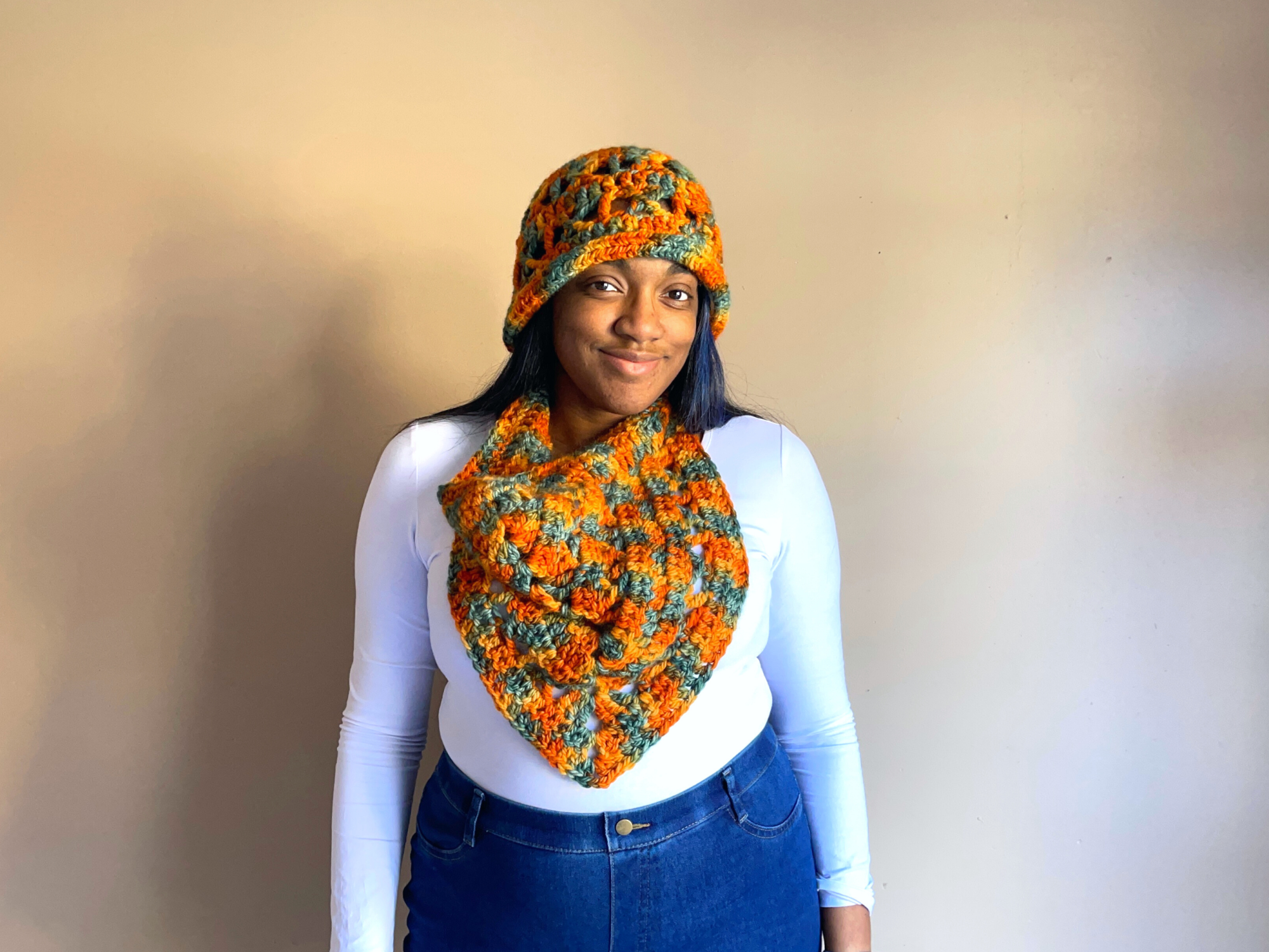 An image of a Black woman wearing a cloche and cowl that hangs down towards the waist. The hat and cowl are made from a chunky yarn that is shades of orange, green and yellow.