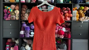 A bright orange crochet top hanging in front of an organized yarn shelf filled with colorful skeins, representing modern, stylish crochet garments made with vibrant and trendy designs