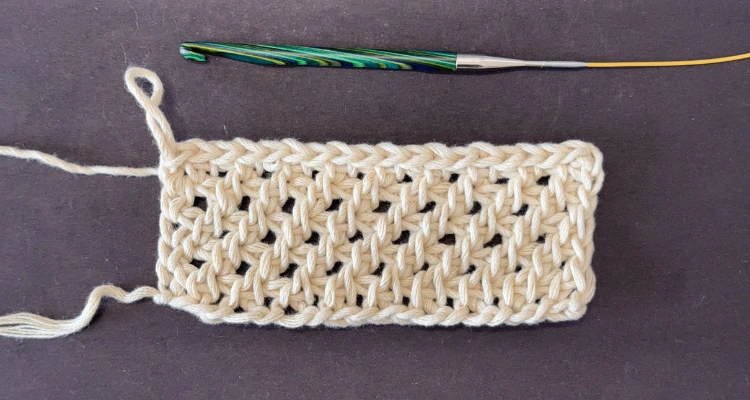 A tunisian crochet tutorial image for the clover stitch. The swatch is made in a natural color, bulky weight cotton yarn that shows the subtle lace and right leaning vertical bars.