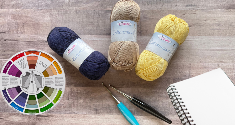 Flat lay of yarn skeins in navy, beige, and yellow with a color wheel, crochet hooks, and a notebook, showing how to choose colors for a crochet project.