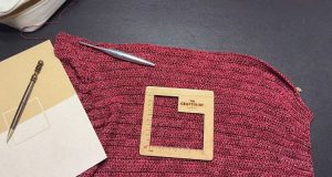 A partially completed piece of red crochet fabric lies on a dark surface, with a silver crochet hook resting on top. A wooden gauge tool labeled "The Craftivist" sits on the fabric to measure stitch size, while a wood-handled pen and sketchpad are placed to the left to check project size.