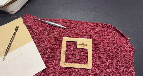 A partially completed piece of red crochet fabric lies on a dark surface, with a silver crochet hook resting on top. A wooden gauge tool labeled "The Craftivist" sits on the fabric to measure stitch size, while a wood-handled pen and sketchpad are placed to the left to check project size.