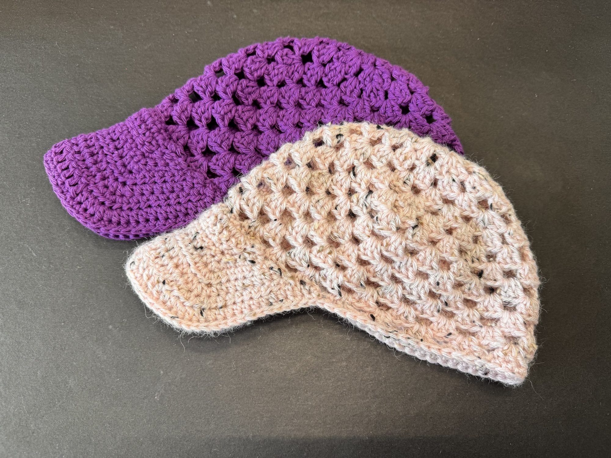 Two handmade crochet baseball caps are displayed on a dark surface. One cap is a solid purple color with a tightly stitched brim, and the other is a speckled pale pink with a similar crocheted texture and structure.
