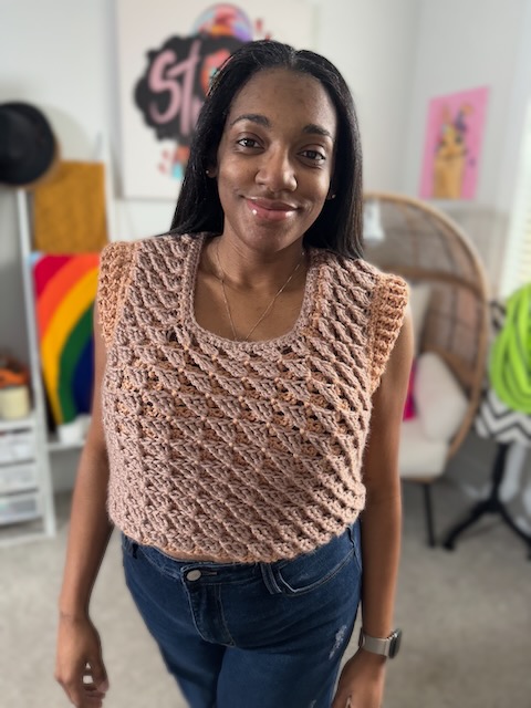 A woman standing indoors and smiling while wearing a handmade crochet lace top in a soft brown yarn with a diamond stitch pattern. She is dressed casually with jeans, and colorful artwork and decor are visible in the background.