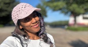 A woman smiling outdoors on a sunny day, wearing a light pink crochet baseball hat with a short brim and lace-style stitching. The background shows trees and a clear blue sky slightly out of focus.