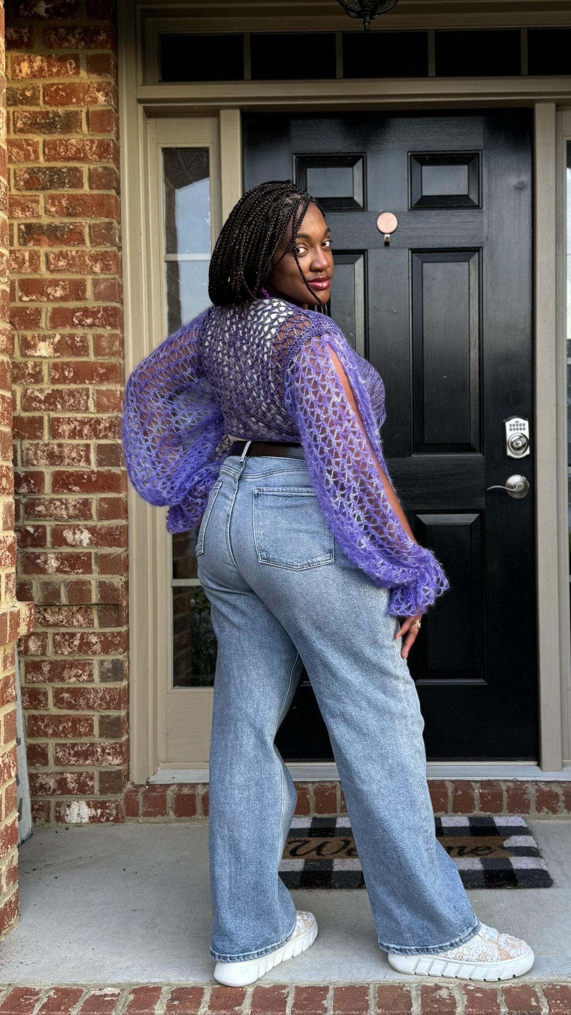 An African American woman standing with her back towards the viewer and the right leg out to the side to showcase the back and sleeves of a cropped bolero. The sleeves are voluminous from the lace weight yarn and features the same stitches as the rectangular crochet shawl pattern.