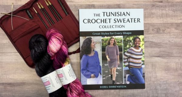 Flat lay of a Tunisian crochet pattern book review setup featuring the book titled "The Tunisian Crochet Sweater Collection Great Styles for Every Shape by Dora Ohrenstein" alongside a set of Tunisian crochet hooks in a red case and two skeins of yarn in black and pink tones on a wooden surface.