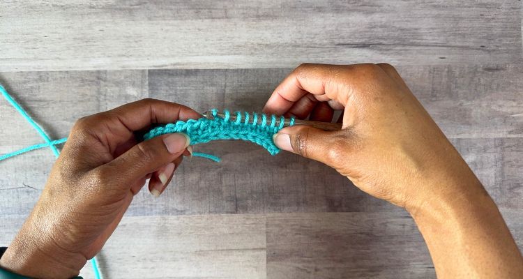 Hands hold a turquoise Tunisian crochet foundation row with multiple loops resting on a crochet hook against a gray wood grain background. The photo demonstrates how the work naturally curves during the forward pass, a common beginner consideration when learning Tunisian crochet tips and techniques.