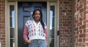 A woman models a cream crochet cardigan with pink and light blue undulating wave motifs, pink sleeves, decorative buttons, and large blue earrings while standing in front of a black door. The close up highlights the cardigan's textured stitches and colorful wave design for the Undulating Waves Cardigan crochet pattern.