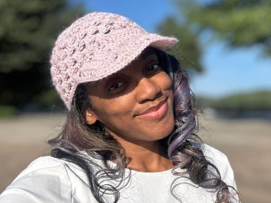 A woman smiling outdoors on a sunny day, wearing a light pink crochet baseball hat with a short brim and lace-style stitching. The background shows trees and a clear blue sky slightly out of focus.