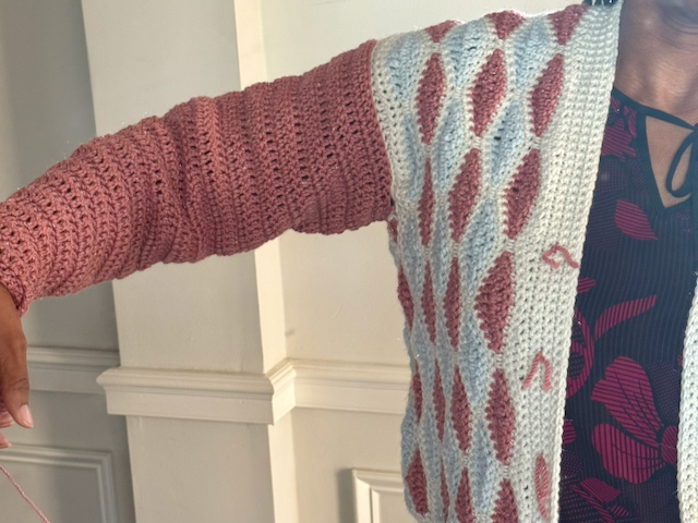 Crocheted cardigan worn with one arm extended, showing a dusty rose sleeve attached to a light blue and burgundy patterned body. The sleeve width and fit are visible from the shoulder to the cuff.